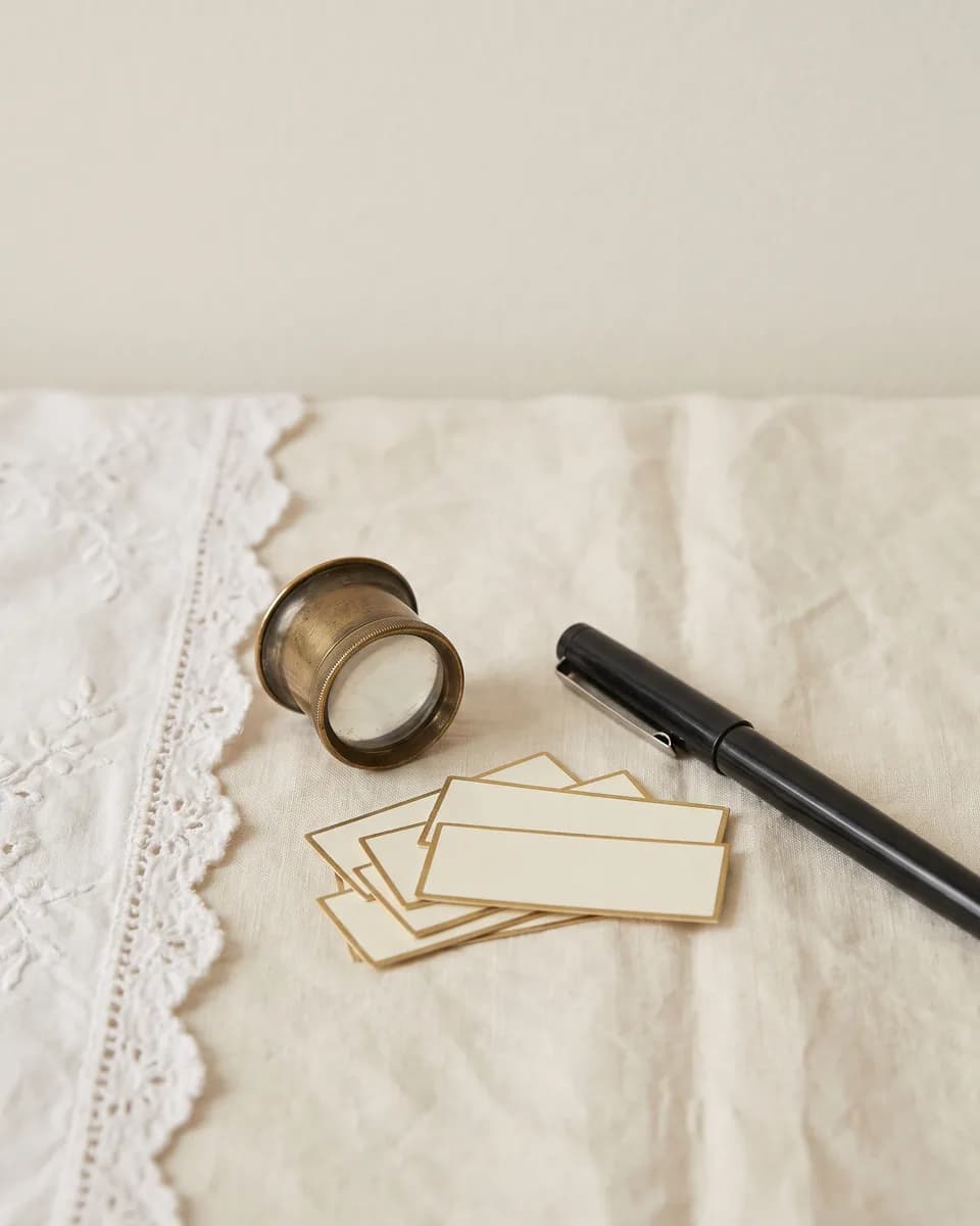 A brass jeweller's loupe, a small stack of blank cream cards with gilt borders and a capped pen on linen beside a lace edge